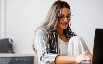 A woman with long hair and glasses is sitting at a table, smiling while working on a laptop. She is wearing a striped shirt over a white top, and appears relaxed in a casual home setting.