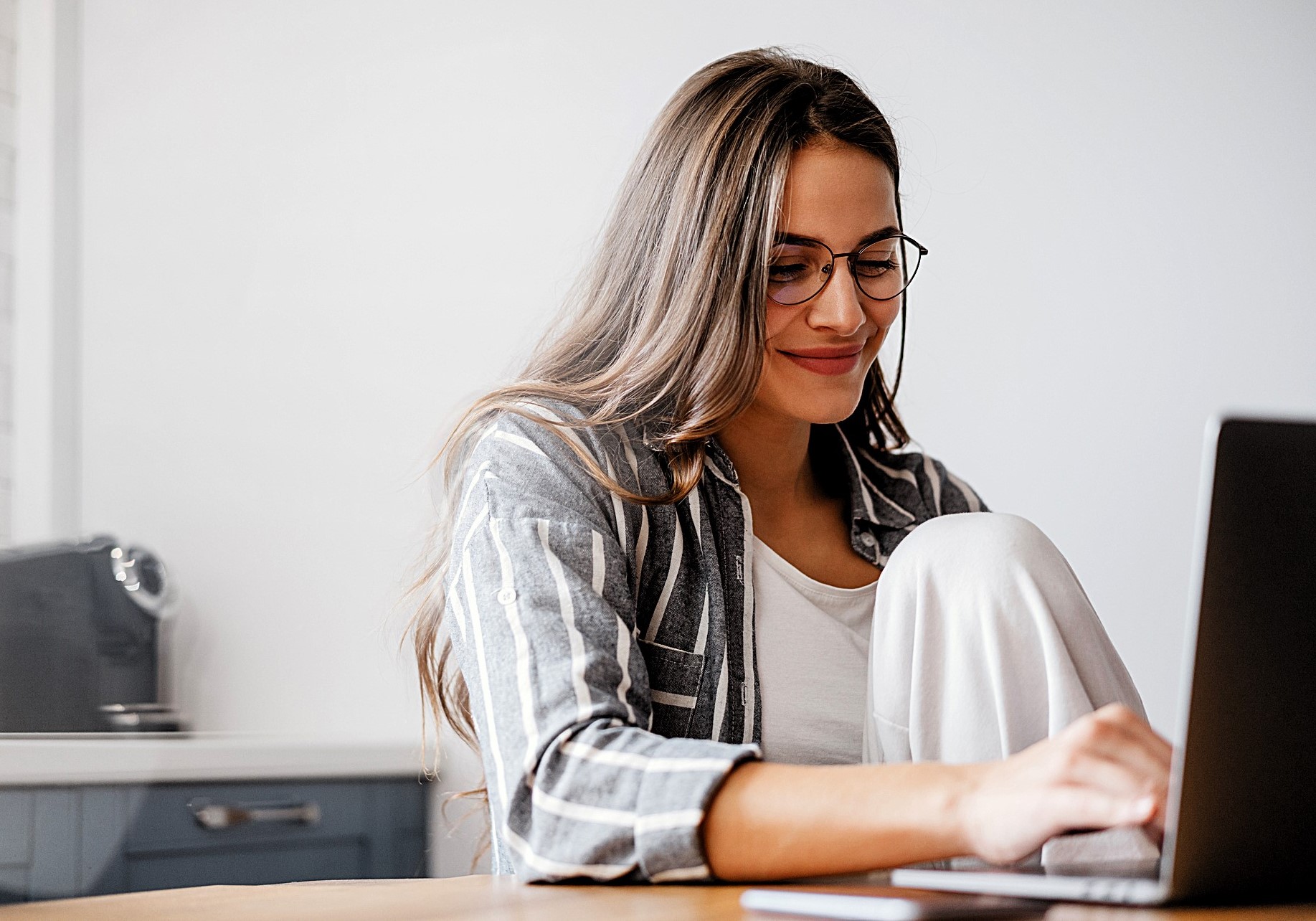 A woman with long hair and glasses is sitting at a table, smiling while working on a laptop. She is wearing a striped shirt over a white top, and appears relaxed in a casual home setting.