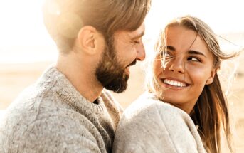 A joyful couple in cozy sweaters smiles at each other, sitting outdoors with a warm, sunlit background. The woman's hair is gently blown by the wind, and both appear relaxed and happy in the soft, natural light.