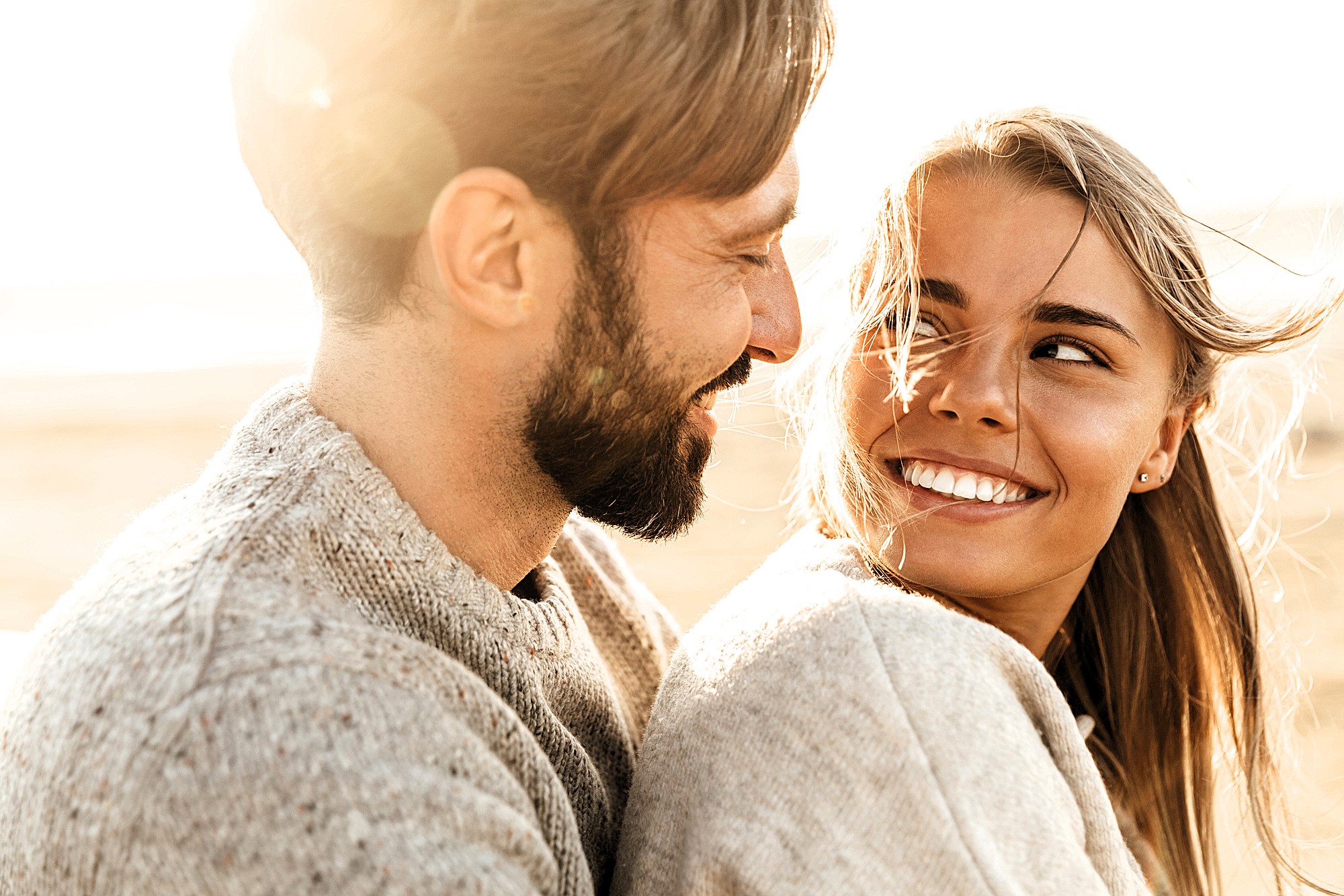 A joyful couple in cozy sweaters smiles at each other, sitting outdoors with a warm, sunlit background. The woman's hair is gently blown by the wind, and both appear relaxed and happy in the soft, natural light.