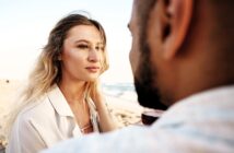 A woman with long blonde hair gazes softly at a person holding a wine glass on the beach. The ocean is visible in the background, and both are sitting closely, creating an intimate and serene atmosphere.