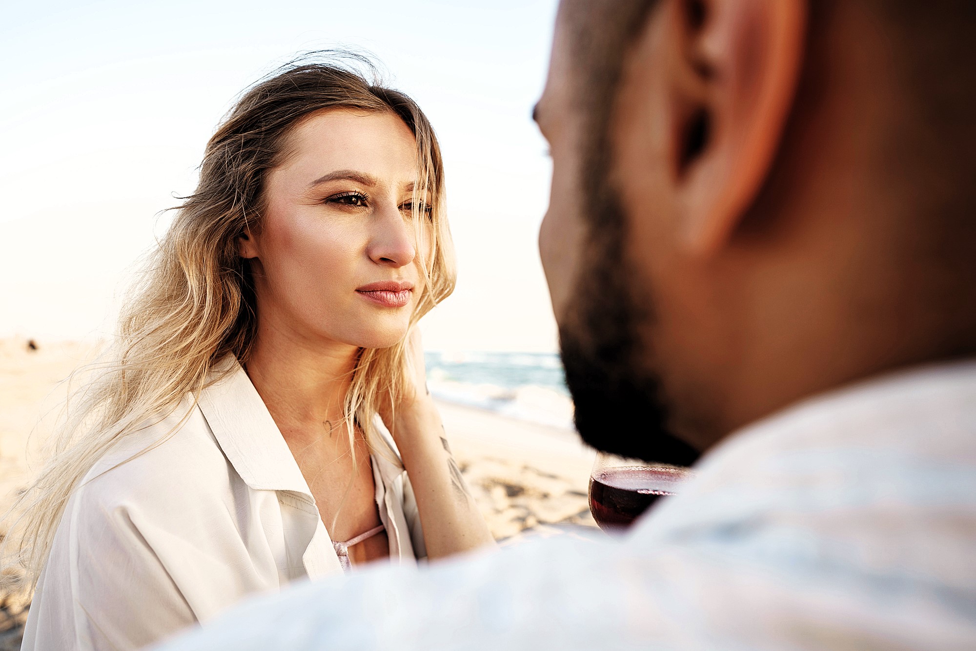 A woman with long blonde hair gazes softly at a person holding a wine glass on the beach. The ocean is visible in the background, and both are sitting closely, creating an intimate and serene atmosphere.