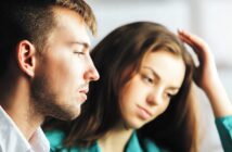 A man and a woman are sitting close together, both gazing thoughtfully into the distance. The man, in a white shirt, is in the foreground, while the woman, with long hair and a green top, is slightly out of focus in the background.