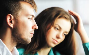 A man and a woman are sitting close together, both gazing thoughtfully into the distance. The man, in a white shirt, is in the foreground, while the woman, with long hair and a green top, is slightly out of focus in the background.