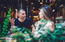 A man and woman sit at an outdoor café table, engaged in a lively conversation. The man gestures with his hand, and the woman listens. The scene is framed by greenery in the foreground, and warm lights create a cozy atmosphere.