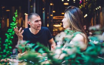 A man and woman sit at an outdoor café table, engaged in a lively conversation. The man gestures with his hand, and the woman listens. The scene is framed by greenery in the foreground, and warm lights create a cozy atmosphere.