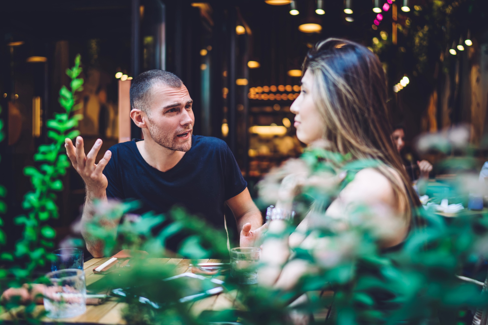 A man and woman sit at an outdoor café table, engaged in a lively conversation. The man gestures with his hand, and the woman listens. The scene is framed by greenery in the foreground, and warm lights create a cozy atmosphere.