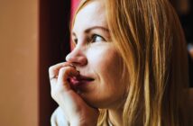 A person with light hair gazes thoughtfully out of a window, resting their chin on their hand. Warm lighting highlights their serene expression.