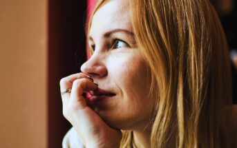 A person with light hair gazes thoughtfully out of a window, resting their chin on their hand. Warm lighting highlights their serene expression.