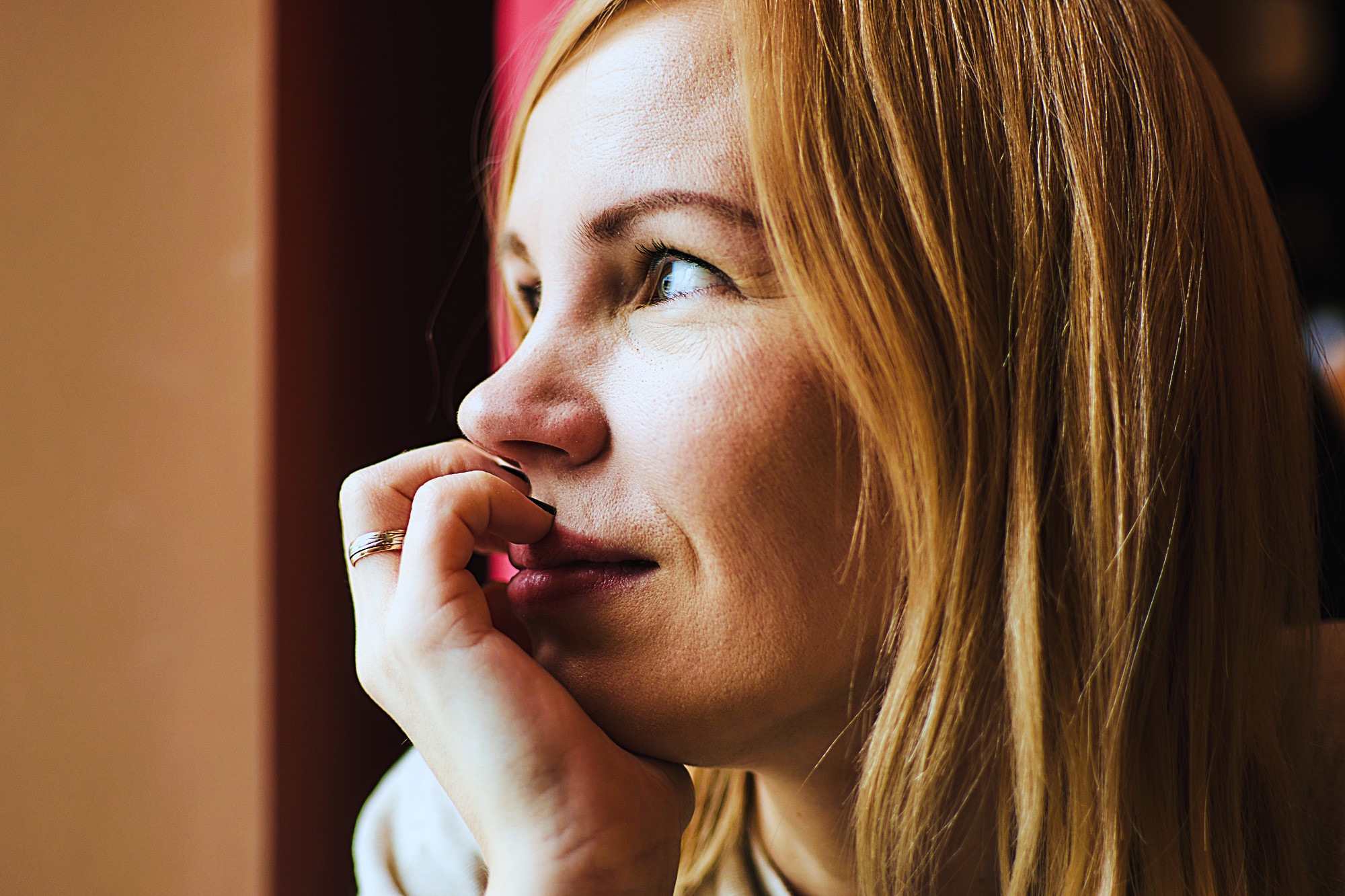 A person with light hair gazes thoughtfully out of a window, resting their chin on their hand. Warm lighting highlights their serene expression.