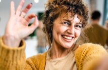 A person with curly hair smiles widely, holding up an "OK" hand gesture. They are wearing a mustard-colored sweater and a beige top. The background is blurred, suggesting an indoor setting with soft lighting.