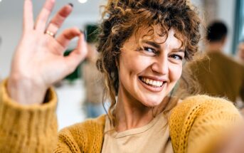 A person with curly hair smiles widely, holding up an "OK" hand gesture. They are wearing a mustard-colored sweater and a beige top. The background is blurred, suggesting an indoor setting with soft lighting.