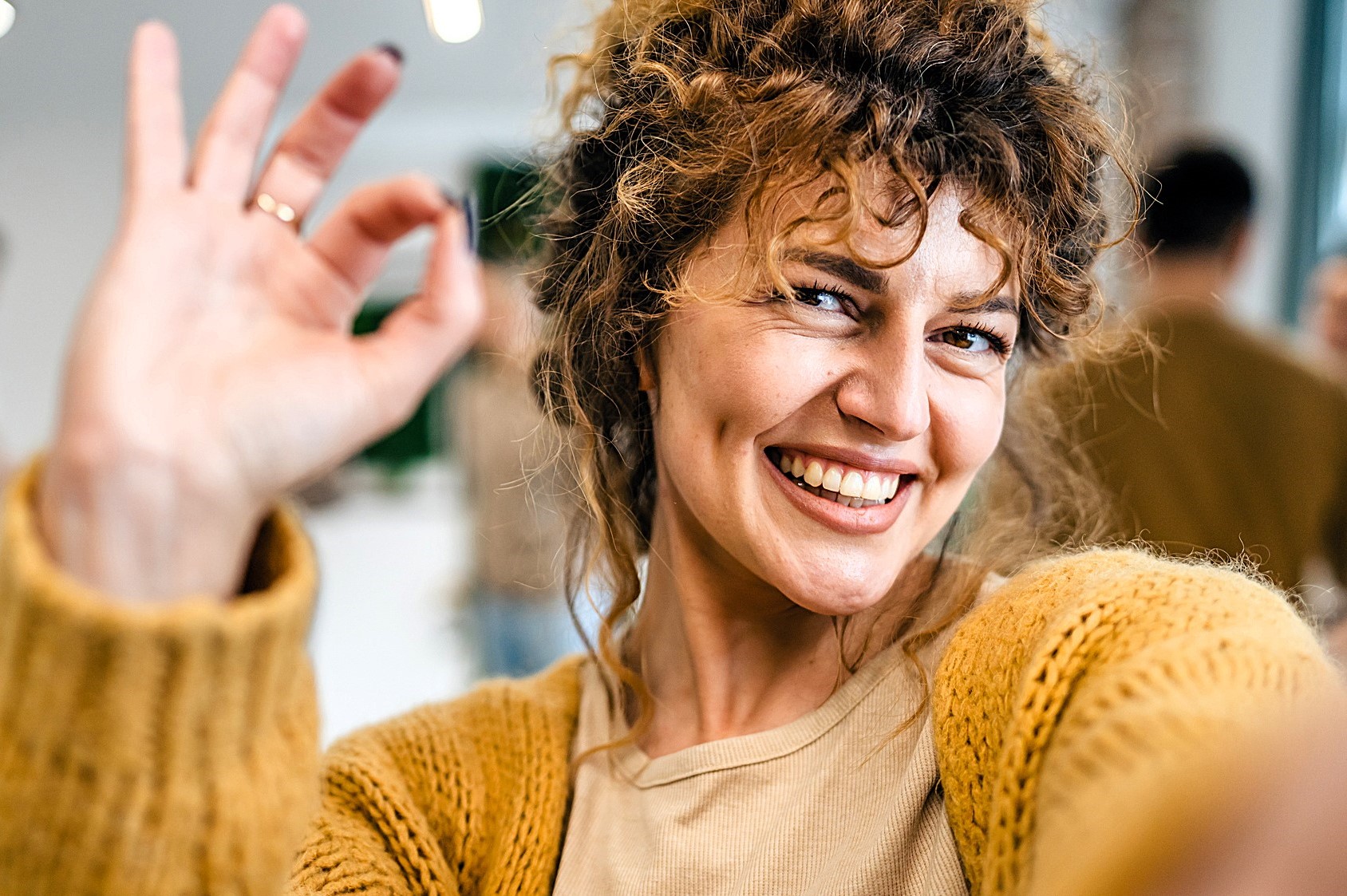 A person with curly hair smiles widely, holding up an "OK" hand gesture. They are wearing a mustard-colored sweater and a beige top. The background is blurred, suggesting an indoor setting with soft lighting.