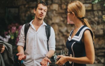 A man and a woman stand outdoors, engaged in conversation. The man gestures with his hands while talking, wearing a white shirt and backpack. The woman, in a blue dress with white accents, holds a cup. They are in a stone-walled setting.