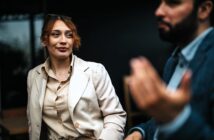 A woman in a beige blazer and blouse looks attentively at a man gesturing with his hand. They appear to be having a conversation in a dimly lit setting. The focus is on the woman, while the man is blurred in the foreground.