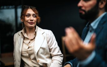 A woman in a beige blazer and blouse looks attentively at a man gesturing with his hand. They appear to be having a conversation in a dimly lit setting. The focus is on the woman, while the man is blurred in the foreground.