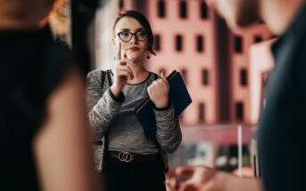 A woman wearing glasses and holding a notebook gestures with her finger, appearing to make a point during a conversation with two blurred individuals in the foreground. She is dressed in a gray top and blazer, standing in an indoor setting.