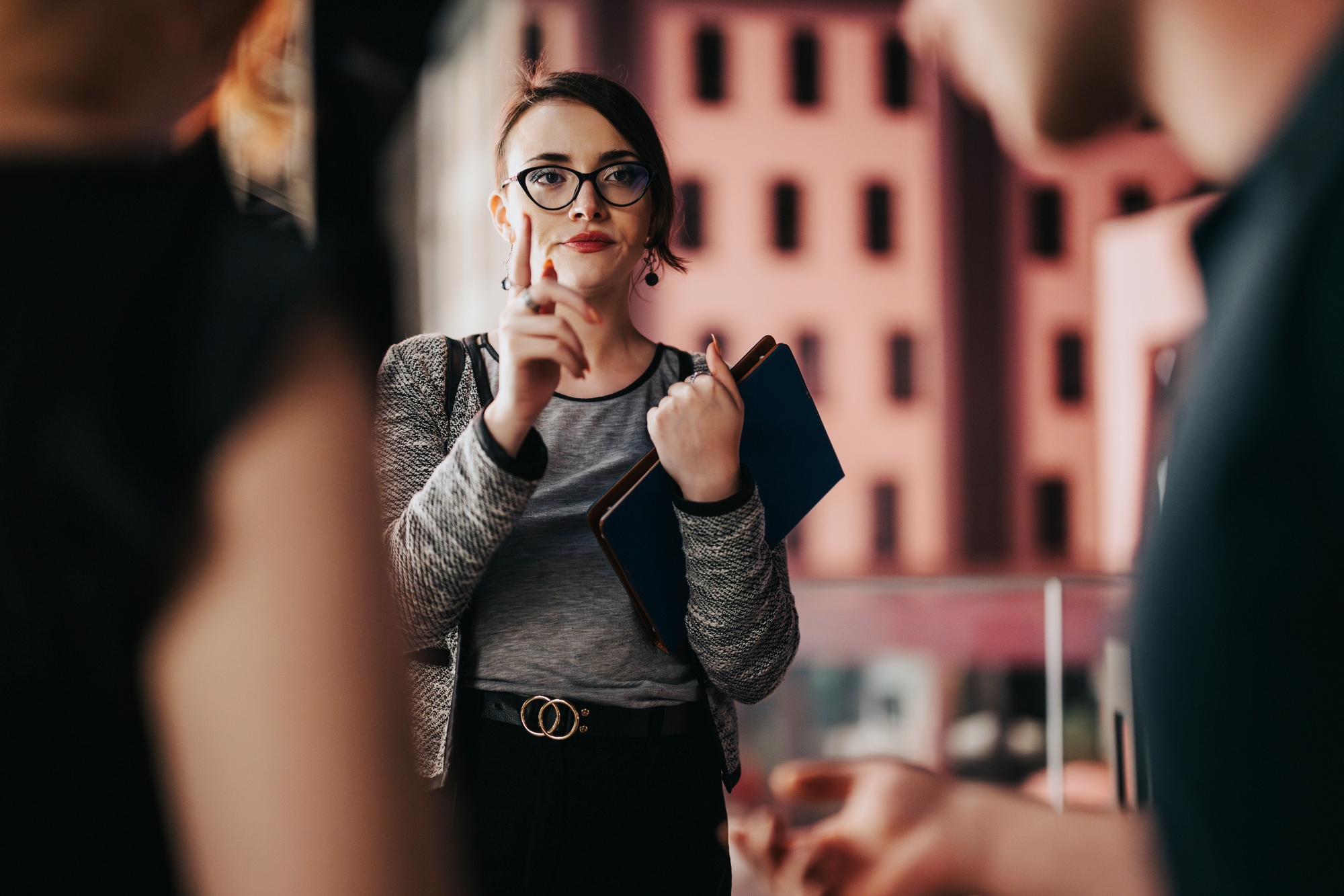 A woman wearing glasses and holding a notebook gestures with her finger, appearing to make a point during a conversation with two blurred individuals in the foreground. She is dressed in a gray top and blazer, standing in an indoor setting.