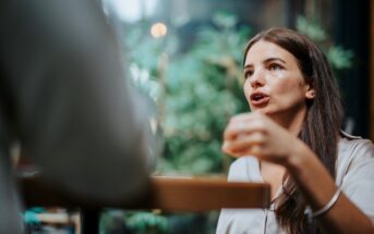 A woman with long brown hair is engaged in conversation, holding a small glass. She is sitting at a table in a bright setting with green plants in the background.