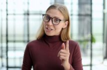 A woman with long blonde hair and black glasses is wearing a maroon turtleneck. She is smiling and pointing her finger, standing in a bright room with blurred windows in the background.