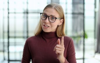 A woman with long blonde hair and black glasses is wearing a maroon turtleneck. She is smiling and pointing her finger, standing in a bright room with blurred windows in the background.