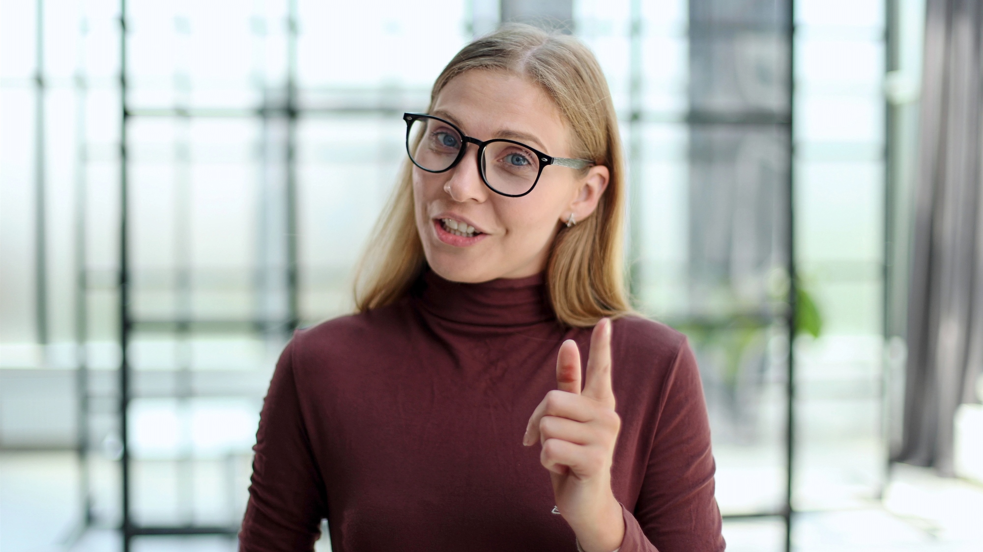 A woman with long blonde hair and black glasses is wearing a maroon turtleneck. She is smiling and pointing her finger, standing in a bright room with blurred windows in the background.