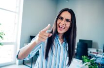 A woman wearing glasses and a striped shirt is standing in an office, pointing forward with an expressive face, possibly angry or emphasizing a point. The room has a large window and a desk with plants and office supplies.