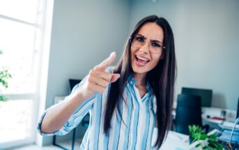 A woman wearing glasses and a striped shirt is standing in an office, pointing forward with an expressive face, possibly angry or emphasizing a point. The room has a large window and a desk with plants and office supplies.