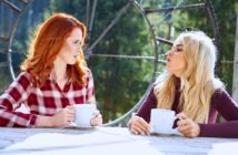 Two women sit at an outdoor table with large ceramic mugs. One woman with red hair wears a plaid shirt, and the other with blonde hair wears a dark top. They are engaged in conversation, with a forest backdrop and large metal wheel structure behind them.