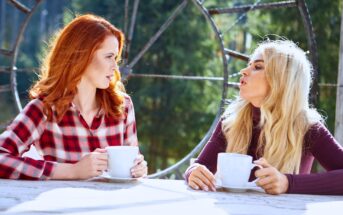 Two women sit at an outdoor table with large ceramic mugs. One woman with red hair wears a plaid shirt, and the other with blonde hair wears a dark top. They are engaged in conversation, with a forest backdrop and large metal wheel structure behind them.