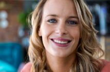 A smiling person with long blonde hair and blue eyes is looking at the camera. The background is blurred with hints of greenery and indoor decor.