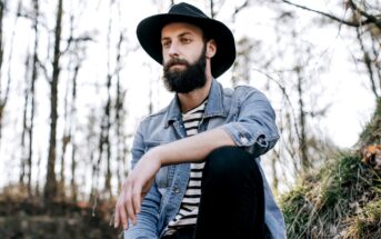 A bearded man wearing a wide-brimmed hat, denim jacket, and striped shirt sits on a grassy hillside in a forest. He gazes thoughtfully into the distance, surrounded by tall, leafless trees under a bright sky.