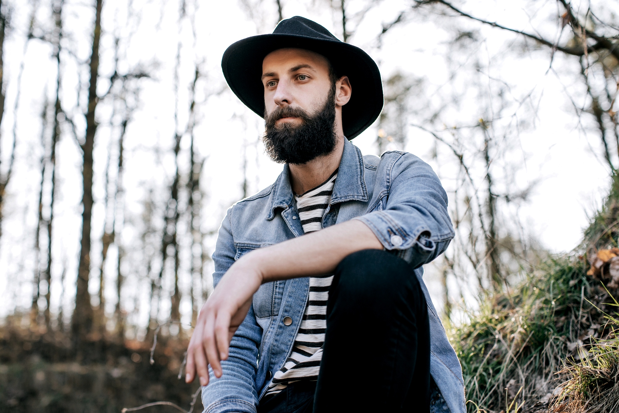 A bearded man wearing a wide-brimmed hat, denim jacket, and striped shirt sits on a grassy hillside in a forest. He gazes thoughtfully into the distance, surrounded by tall, leafless trees under a bright sky.