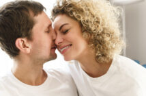 A couple wearing white shirts is smiling affectionately with their noses touching. The man has short brown hair, and the woman has curly blonde hair. They appear happy and are facing each other closely, suggesting a moment of intimacy.