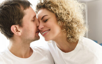 A couple wearing white shirts is smiling affectionately with their noses touching. The man has short brown hair, and the woman has curly blonde hair. They appear happy and are facing each other closely, suggesting a moment of intimacy.