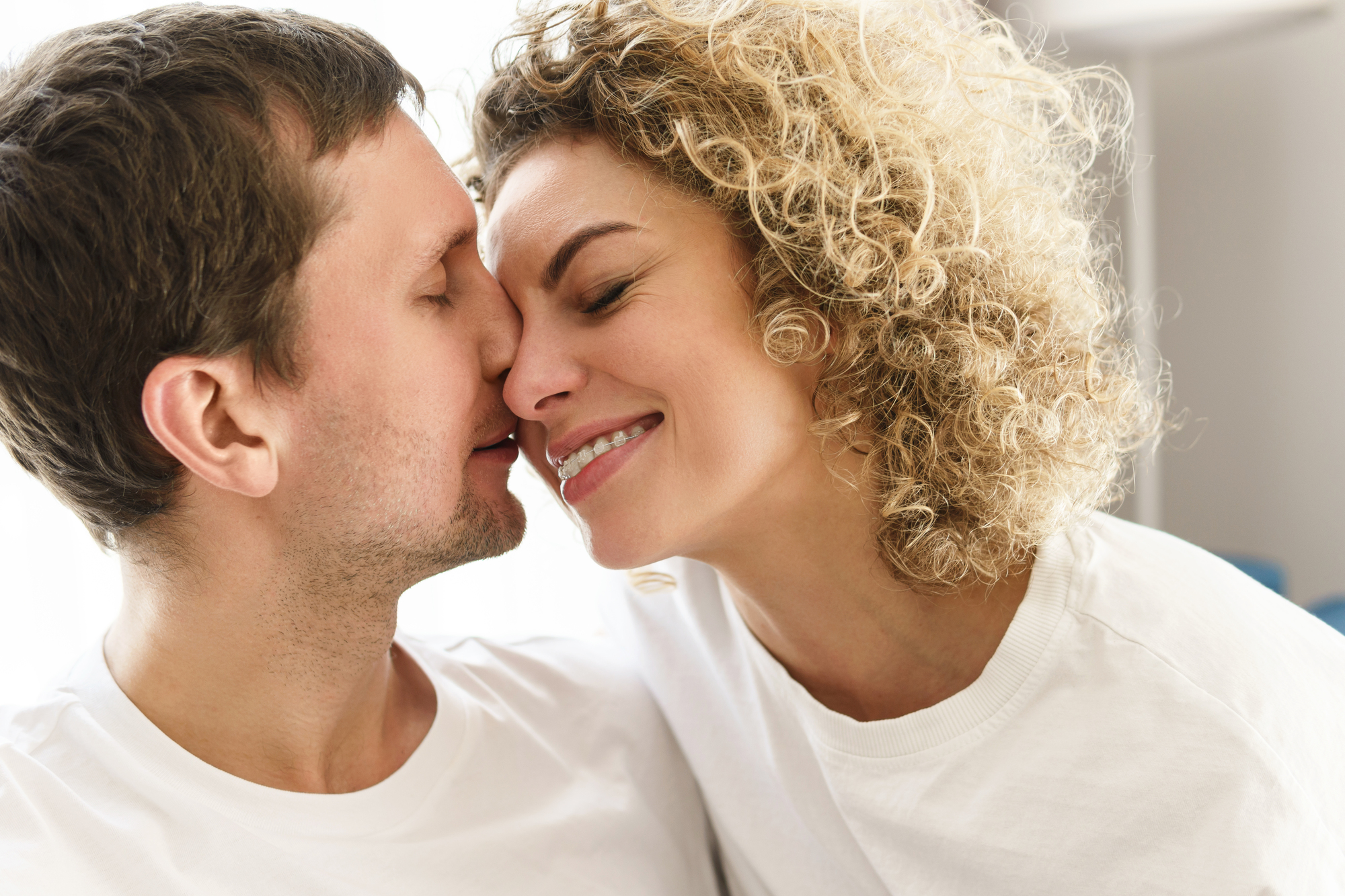 A couple wearing white shirts is smiling affectionately with their noses touching. The man has short brown hair, and the woman has curly blonde hair. They appear happy and are facing each other closely, suggesting a moment of intimacy.