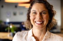 A woman with short brown hair smiles warmly at the camera. She is wearing a white shirt with small black dots. The background is softly blurred, showing a cozy indoor setting with warm lighting.