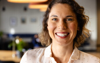 A woman with short brown hair smiles warmly at the camera. She is wearing a white shirt with small black dots. The background is softly blurred, showing a cozy indoor setting with warm lighting.