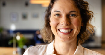 A woman with short brown hair smiles warmly at the camera. She is wearing a white shirt with small black dots. The background is softly blurred, showing a cozy indoor setting with warm lighting.