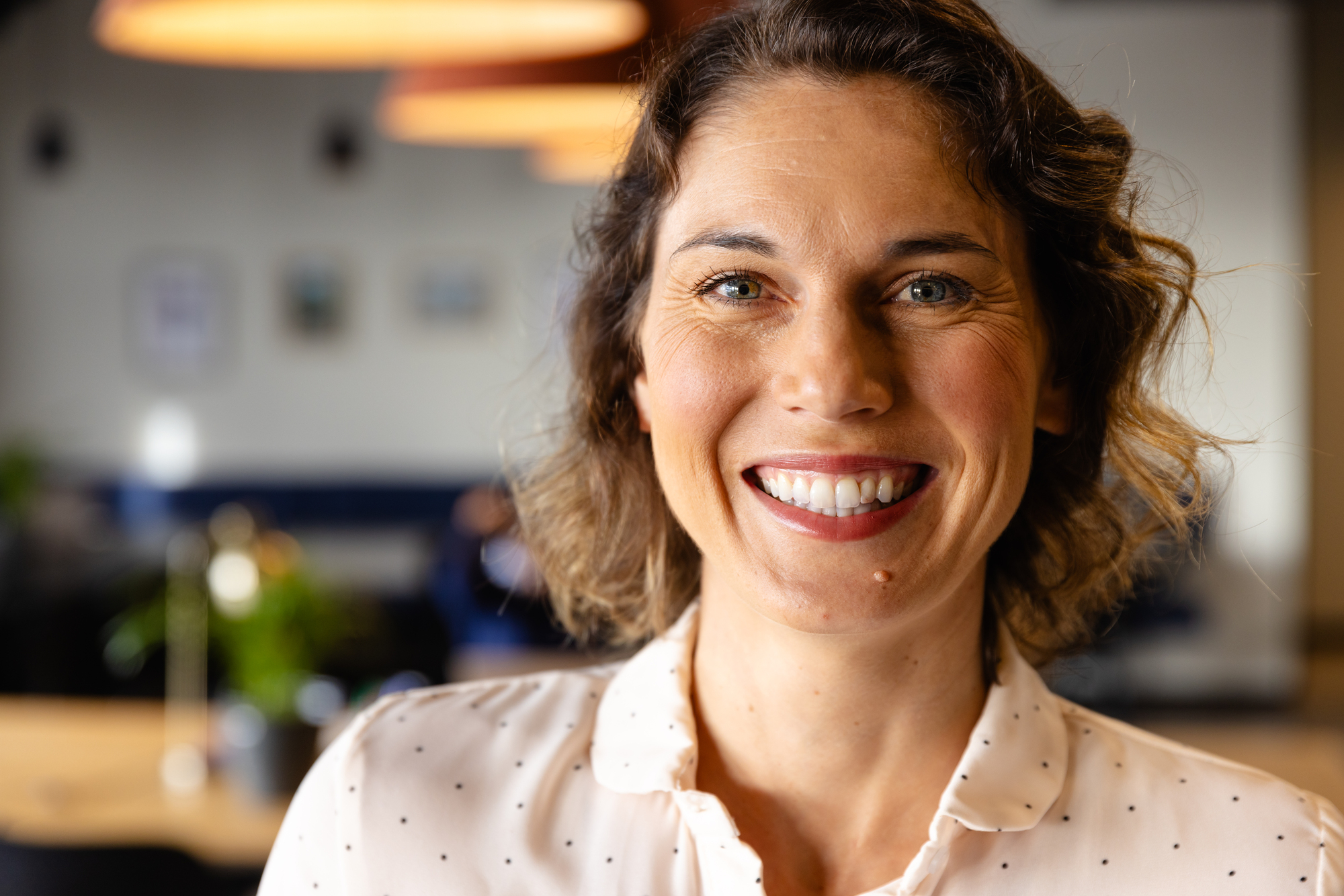 A woman with short brown hair smiles warmly at the camera. She is wearing a white shirt with small black dots. The background is softly blurred, showing a cozy indoor setting with warm lighting.