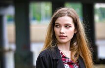 A woman with long hair and a plaid shirt looks to the side with a thoughtful expression. She is outdoors, with blurred columns and greenery in the background.