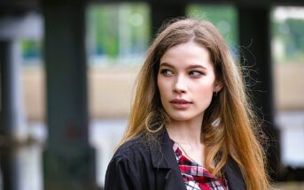 A woman with long hair and a plaid shirt looks to the side with a thoughtful expression. She is outdoors, with blurred columns and greenery in the background.