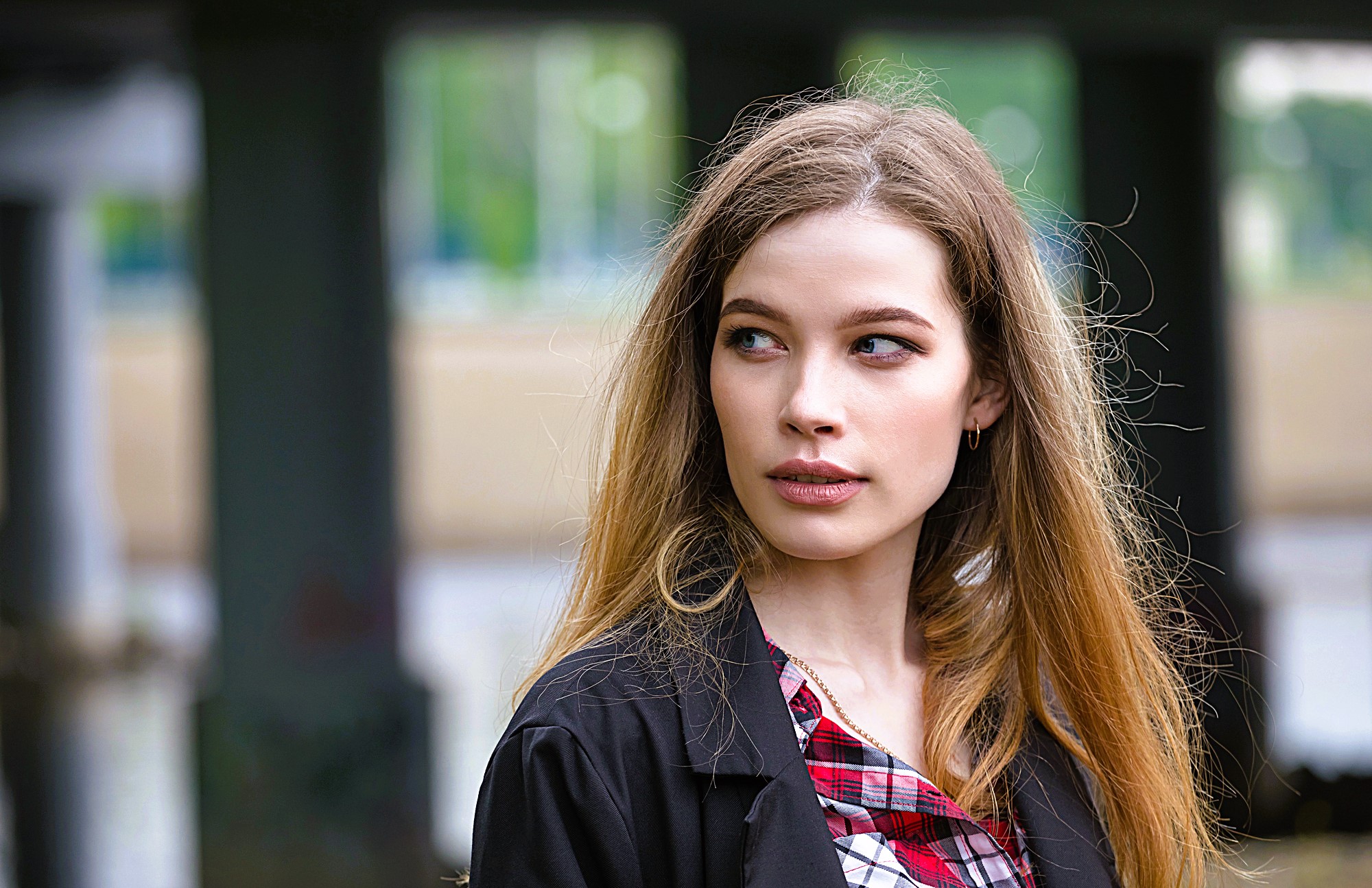 A woman with long hair and a plaid shirt looks to the side with a thoughtful expression. She is outdoors, with blurred columns and greenery in the background.