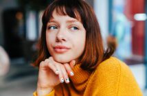 A person with chin-length brown hair wearing a yellow sweater, looking pensively to the side. Their hand is resting on their chin, showing painted nails. The background is softly blurred.