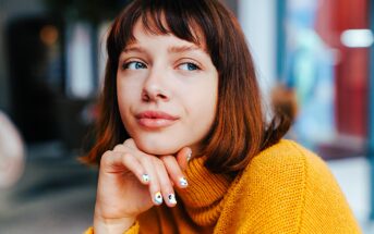 A person with chin-length brown hair wearing a yellow sweater, looking pensively to the side. Their hand is resting on their chin, showing painted nails. The background is softly blurred.