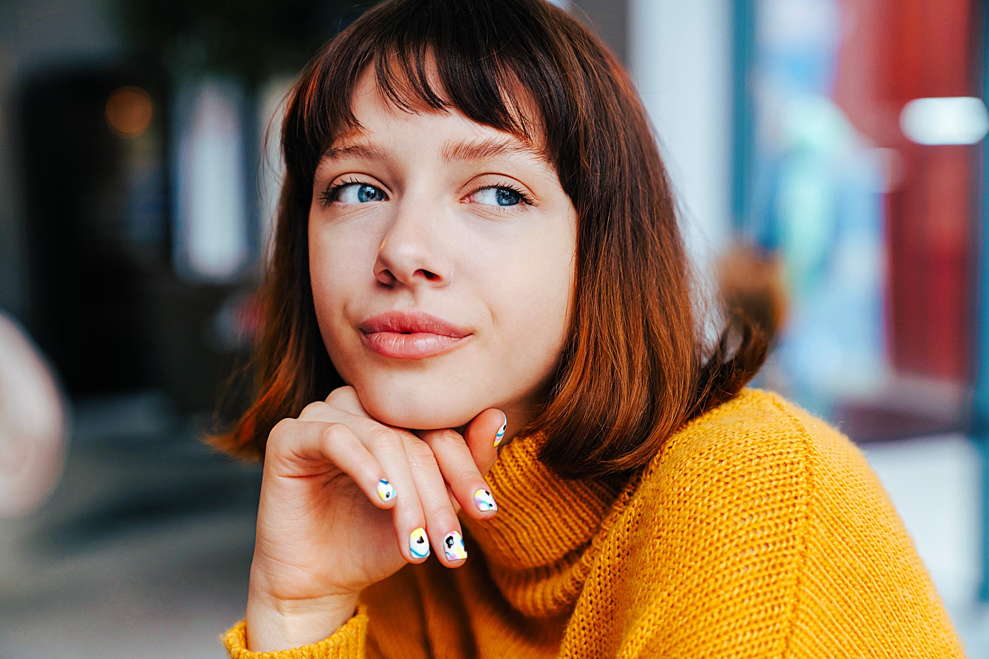 A person with chin-length brown hair wearing a yellow sweater, looking pensively to the side. Their hand is resting on their chin, showing painted nails. The background is softly blurred.