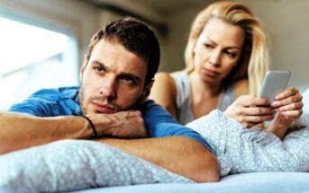 A man and woman lie on a bed, the man looking thoughtfully into the distance while resting his head on his arms. The woman, sitting behind him, holds a smartphone and gazes at him with concern. They are in a softly lit room with a window.