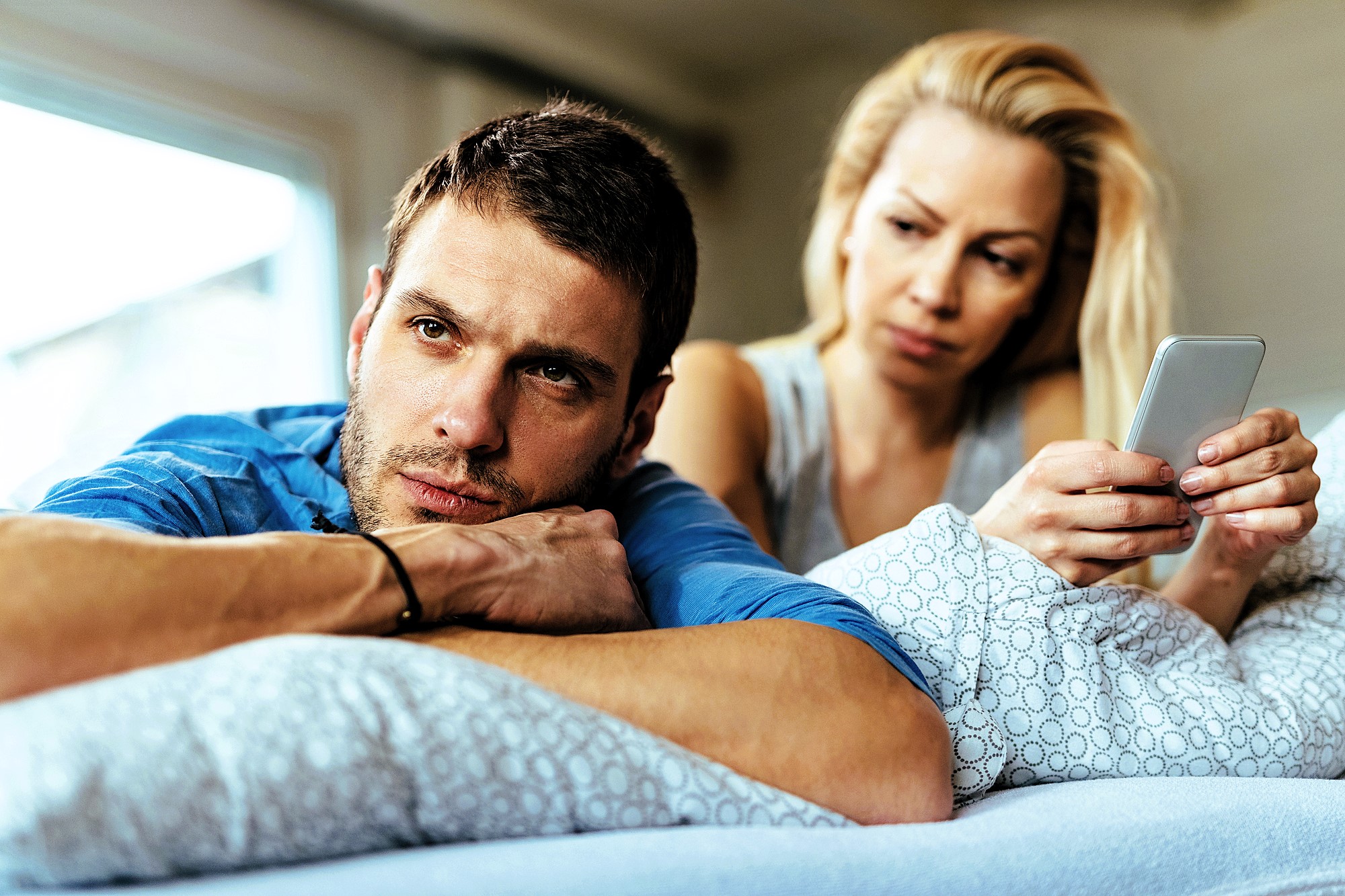 A man and woman lie on a bed, the man looking thoughtfully into the distance while resting his head on his arms. The woman, sitting behind him, holds a smartphone and gazes at him with concern. They are in a softly lit room with a window.