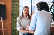 Two people are sitting at a wooden table in a conversation. The woman, wearing a light sweater, appears to be speaking with an expressive gesture, while the man, with curly hair, listens attentively. The setting is a cozy indoor space.