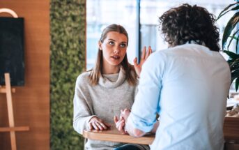 Two people are sitting at a wooden table in a conversation. The woman, wearing a light sweater, appears to be speaking with an expressive gesture, while the man, with curly hair, listens attentively. The setting is a cozy indoor space.